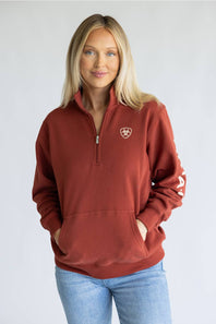 A woman wearing a rust-colored quarter-zip sweatshirt with a small shield logo on the left chest, paired with light blue jeans, standing against a light gray studio background.