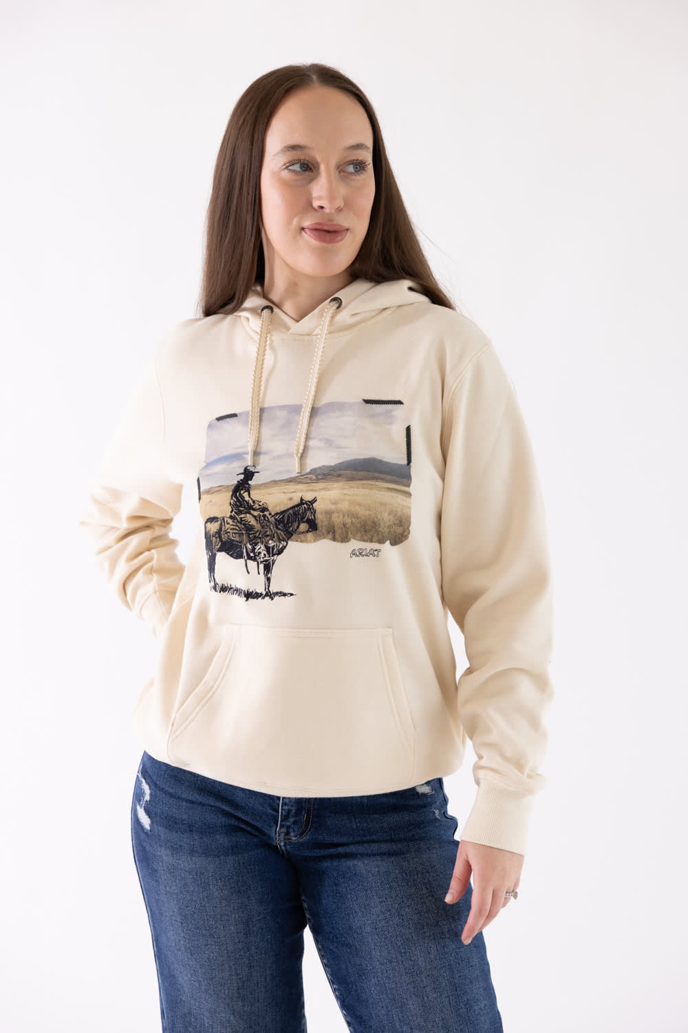 Medium shot of a model wearing the Ariat Wish You Were Here Hoodie in parchment beige, showing the front kangaroo pocket, drawstrings, and vintage western horseback cowboy graphic, paired with dark denim jeans.