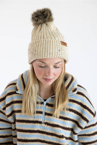 A woman wearing a cream ribbed knit beanie with a brown faux-fur pom-pom and a small brown brand patch, paired with a blue, cream, and brown striped fleece pullover, against a plain white background.