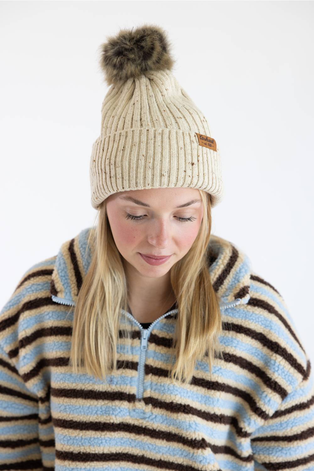 A woman wearing a cream ribbed knit beanie with a brown faux-fur pom-pom and a small brown brand patch, paired with a blue, cream, and brown striped fleece pullover, against a plain white background.