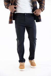 A person wearing slim-fit dark navy pants, styled with a white t-shirt, an open brown and black plaid flannel shirt, and tan suede sneakers, shown from the waist down on a white studio background.