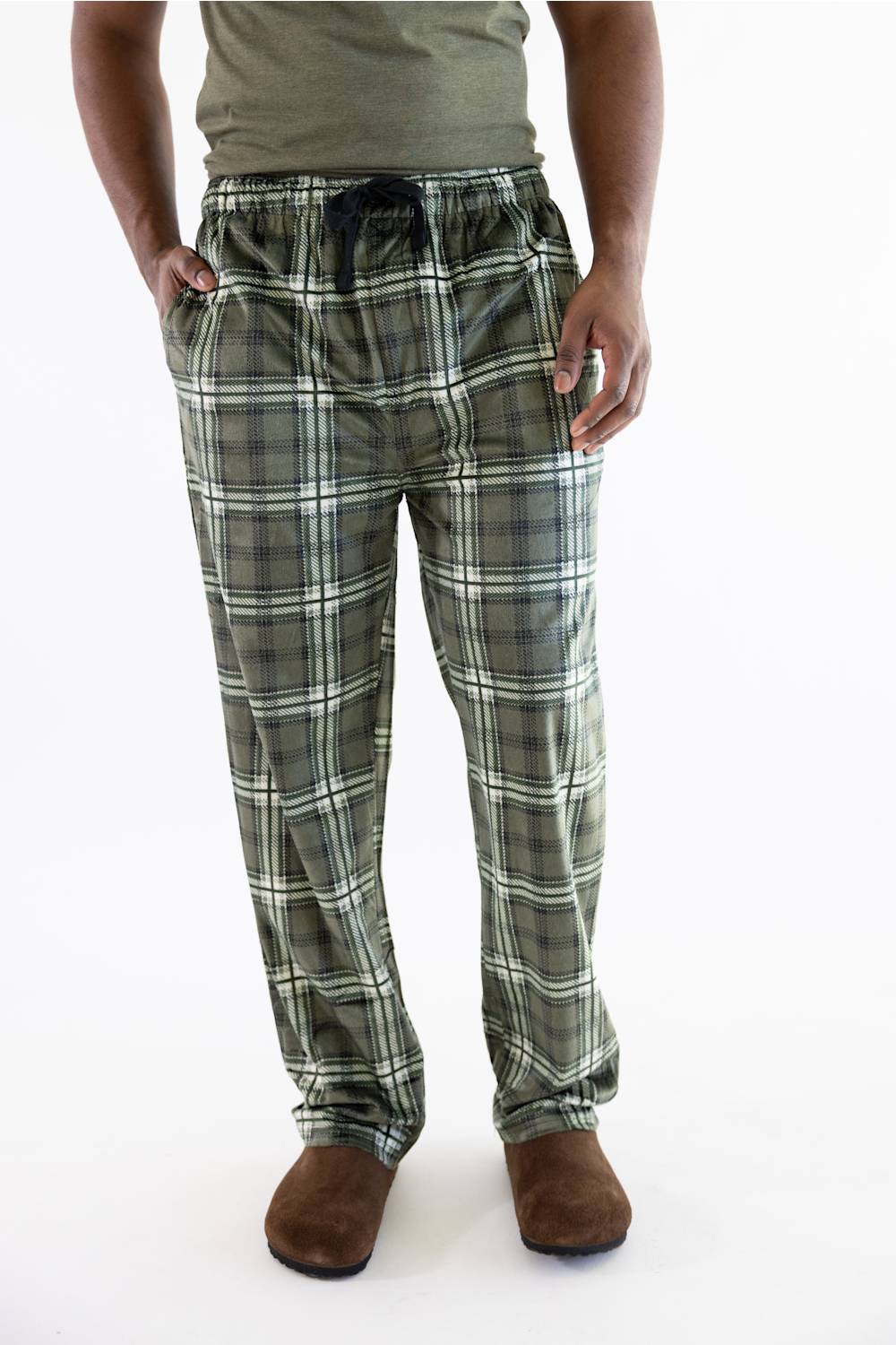 A person wearing plaid lounge pants in olive green, gray, and white, paired with a gray t-shirt and brown slippers, shown from the waist down on a white background.
