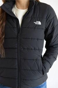 A close-up of a person wearing a black puffer jacket with horizontal quilting, a full front zipper, and a white embroidered 'The North Face' logo on the left chest. The jacket is layered over a cream-colored top.