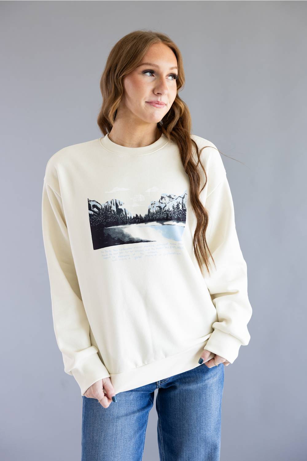 A woman wearing a cream-colored crewneck sweatshirt with a black and white landscape graphic on the front, paired with blue jeans, standing against a solid gray studio background.