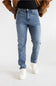 A person wearing light blue slim-fit jeans, styled with a black t-shirt, a brown jacket, and white sneakers, shown from the waist down against a plain white studio background.
