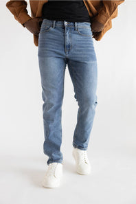 A person wearing light blue slim-fit jeans, styled with a black t-shirt, a brown jacket, and white sneakers, shown from the waist down against a plain white studio background.