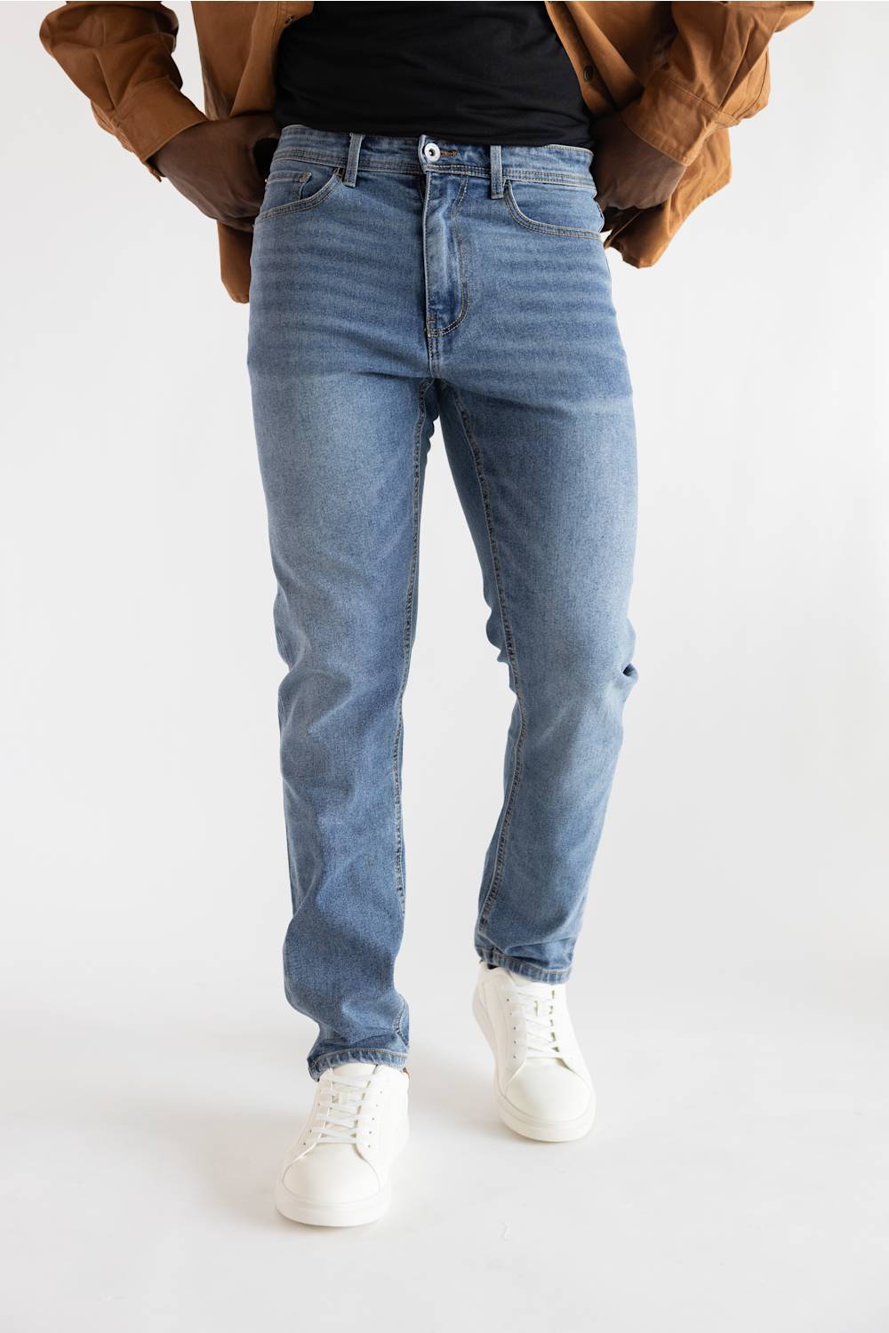 A person wearing light blue slim-fit jeans, styled with a black t-shirt, a brown jacket, and white sneakers, shown from the waist down against a plain white studio background.