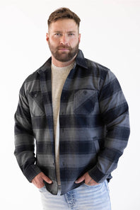 Front, mid-shot of a man modeling the Weatherproof Vintage Lumberjack Plaid Shirt Jacket in Phantom Night (charcoal grey and dark navy plaid) worn open over a beige sweater and light-wash jeans, looking directly at the camera with his hands in his pockets against a white studio background.