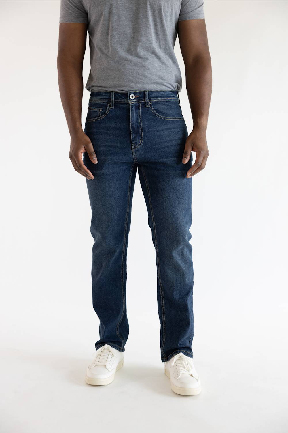 A person wearing dark blue slim-fit jeans, a gray t-shirt, and white sneakers, shown from the waist down against a plain white studio background.