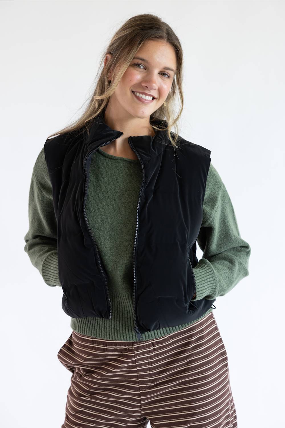 A woman wearing a black zip-up puffer vest over an olive green knit sweater and brown striped pants, photographed against a clean white studio background.