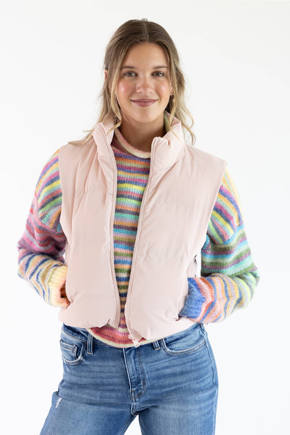 A woman wearing a light pink sleeveless puffer vest over a multicolored striped knit sweater and blue jeans, photographed against a plain white studio background.