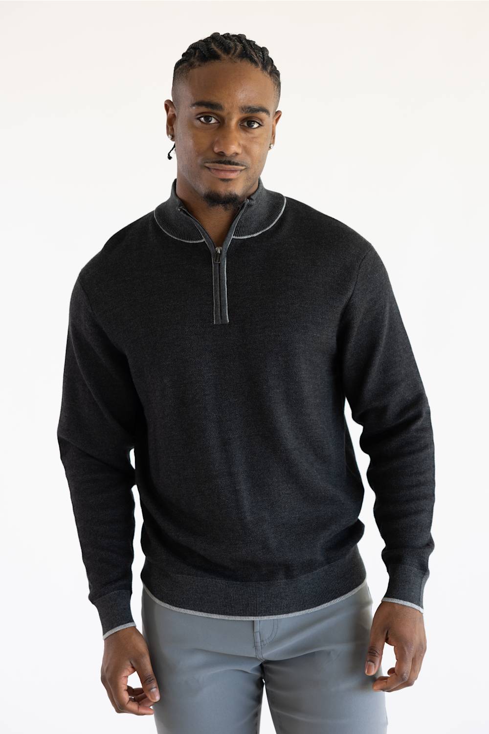 A man wearing a dark charcoal gray quarter-zip sweater with light gray piping details on the collar and cuffs, paired with light gray trousers, against a plain white studio background.