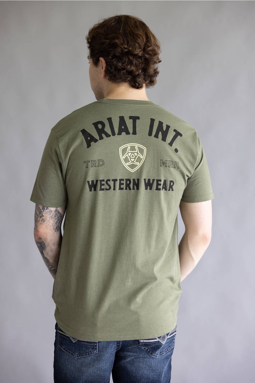 A man viewed from behind wearing an olive green t-shirt with 'ARIAT INT. WESTERN WEAR' printed in black on the back, paired with blue jeans against a gray studio background.