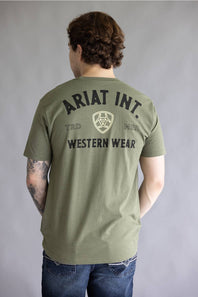 A man viewed from behind wearing an olive green t-shirt with 'ARIAT INT. WESTERN WEAR' printed in black on the back, paired with blue jeans against a gray studio background.