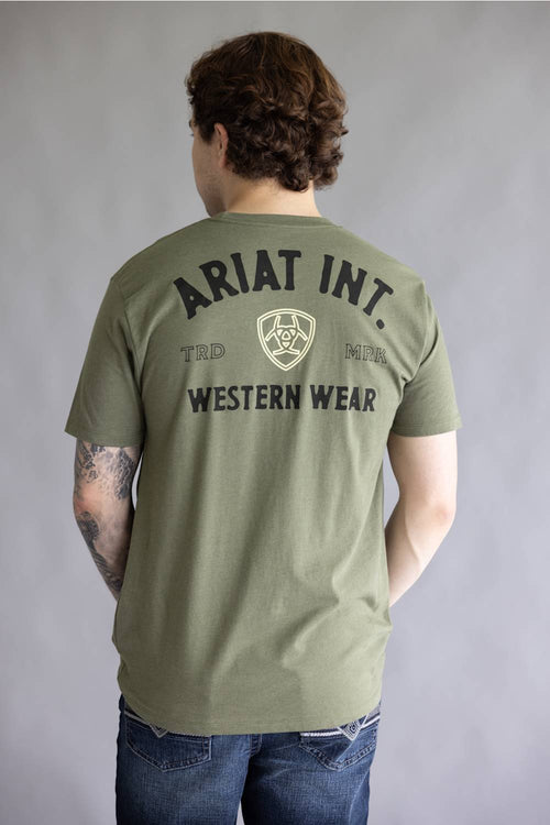 A man viewed from behind wearing an olive green t-shirt with 'ARIAT INT. WESTERN WEAR' printed in black on the back, paired with blue jeans against a gray studio background.