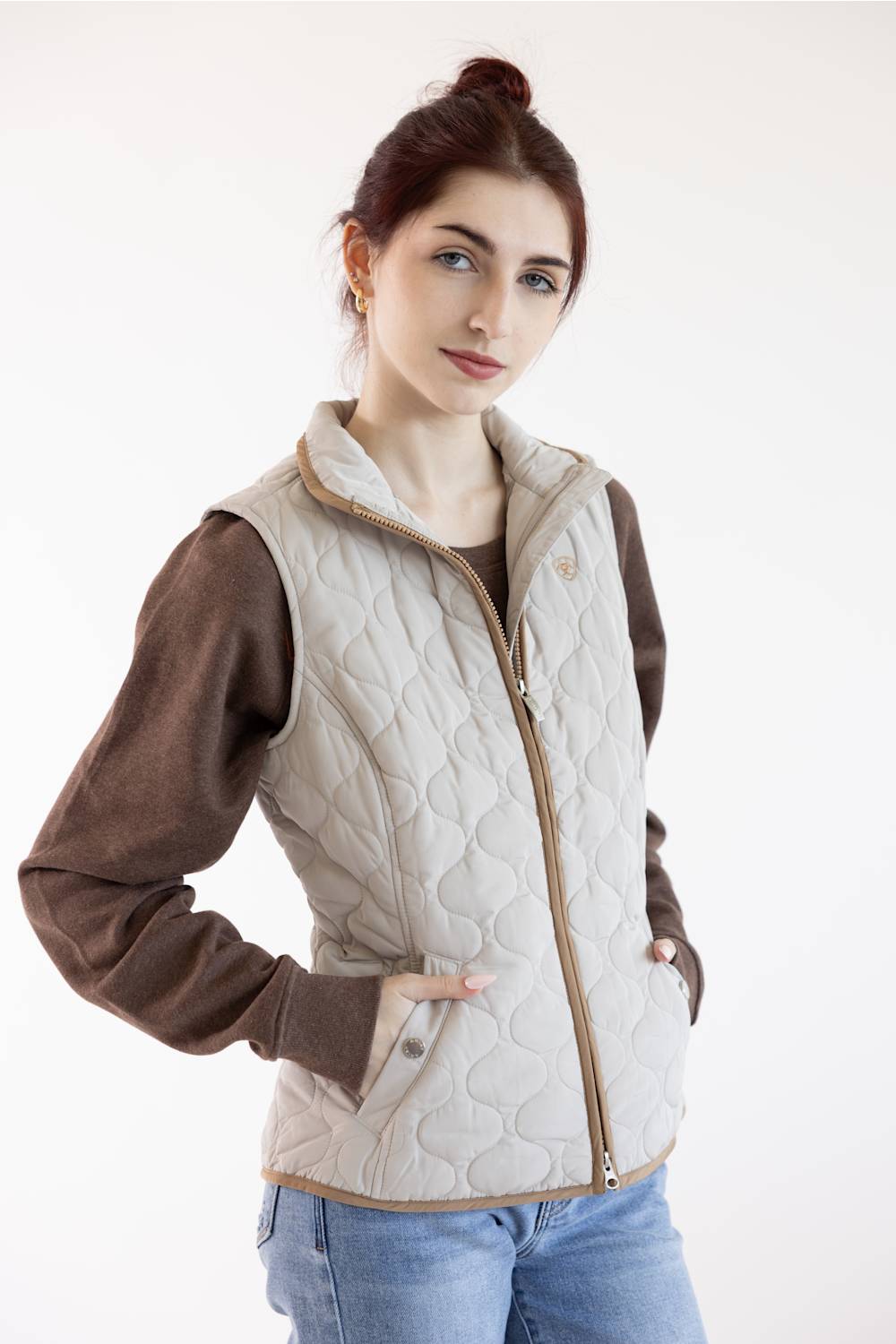 A woman wearing a light beige quilted vest over a brown long-sleeve top and blue jeans, photographed against a plain white studio background.