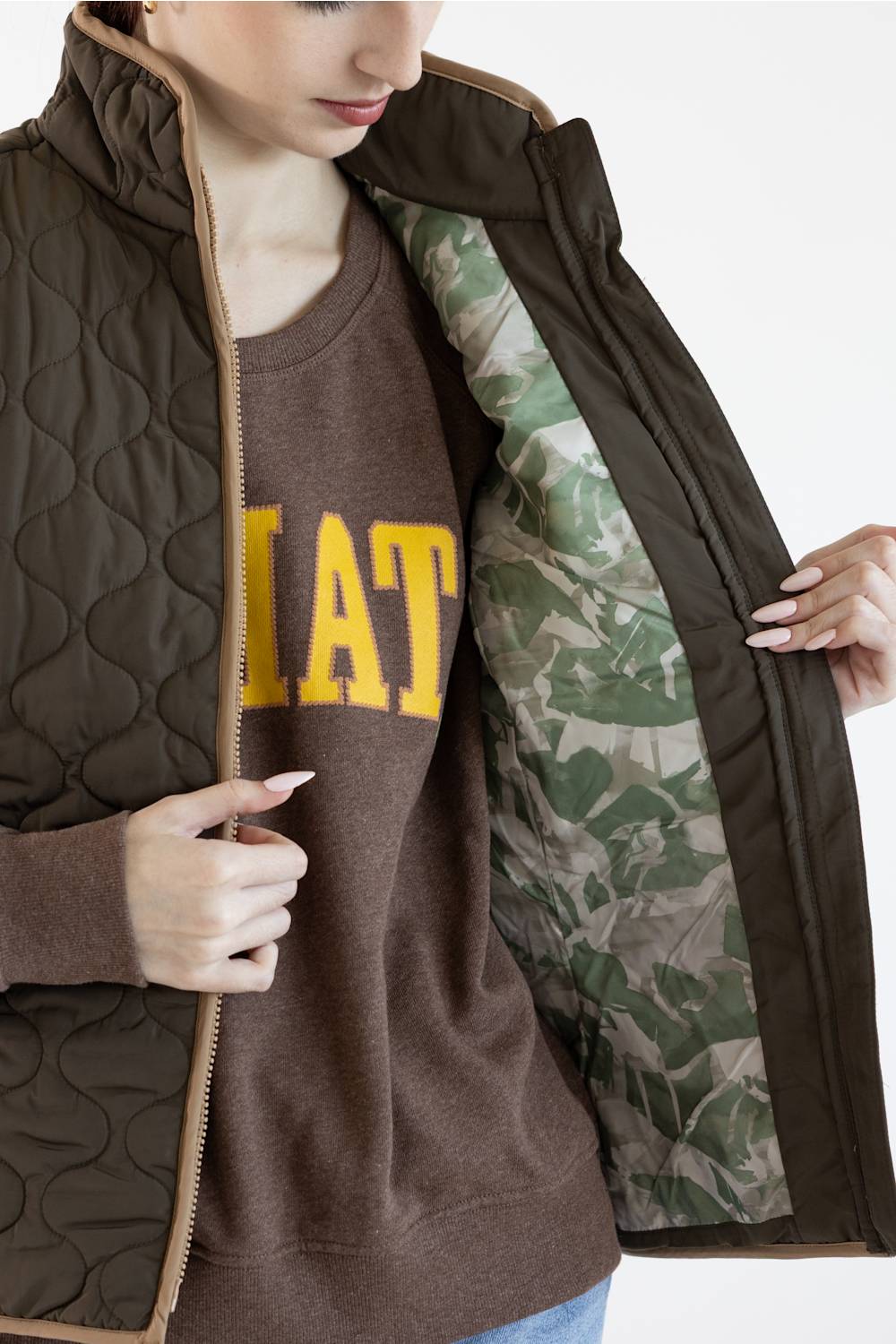 A close-up of a model wearing a dark brown quilted zip-up vest over a brown sweatshirt with yellow lettering, while opening the vest to show a green camouflage patterned lining.