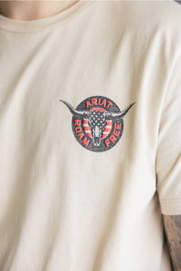 A close-up of a person wearing a cream-colored t-shirt with a circular chest graphic featuring a longhorn skull over an American flag and the words 'ARIAT ROAD FREE'.