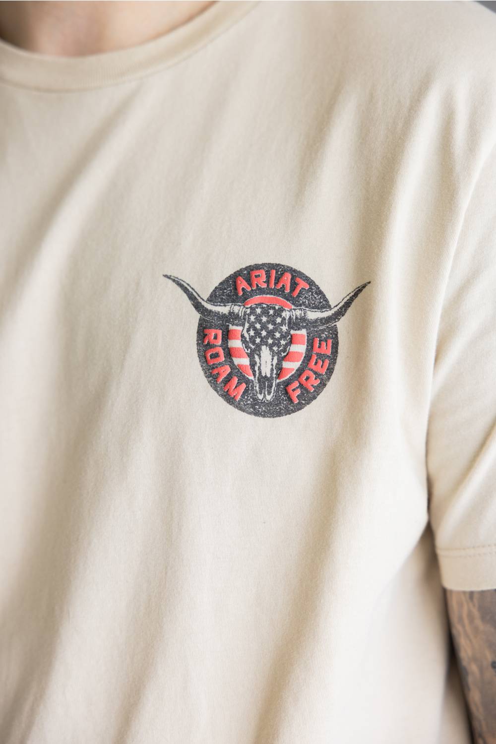 A close-up of a person wearing a cream-colored t-shirt with a circular chest graphic featuring a longhorn skull over an American flag and the words 'ARIAT ROAD FREE'.