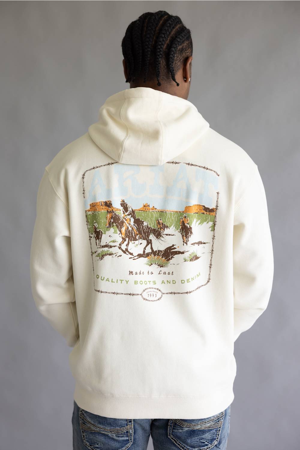 A photo from the back of a man wearing an off-white hoodie featuring a large vintage-style western graphic print with barbed wire, cowboys on horseback, and text reading 'Made to Last Quality Boots and Denim' and 'Since 1993'.
