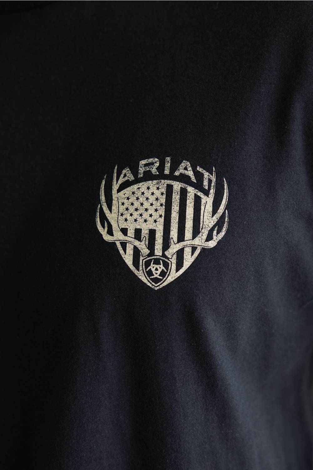 Close-up view of the chest graphic on a black shirt, featuring an American flag shield framed by deer antlers with the Ariat brand name arched above it.