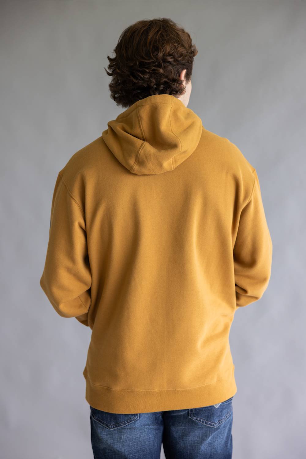 A person viewed from behind wearing a mustard yellow pullover hoodie and blue jeans against a plain gray studio background.