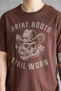A person wearing a brown short-sleeve t-shirt with a distressed light beige graphic of a skull in a cowboy hat and the text 'ARIAT BOOTS' and 'TRAIL WORN'.