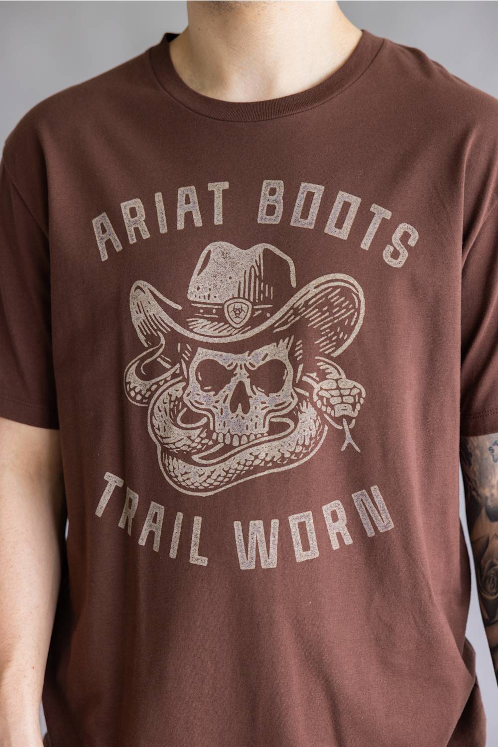 A person wearing a brown short-sleeve t-shirt with a distressed light beige graphic of a skull in a cowboy hat and the text 'ARIAT BOOTS' and 'TRAIL WORN'.