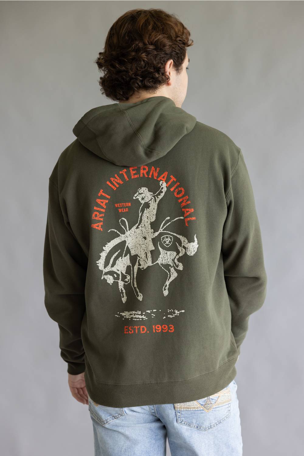 A person viewed from behind wearing an olive green hoodie with a large white and orange graphic on the back, paired with light blue jeans, against a neutral gray studio background.
