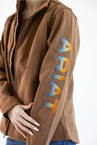 A close-up of a person wearing a brown zip-front jacket with a large colorful striped ARIAT logo printed vertically on the sleeve, paired with blue jeans against a plain white background.