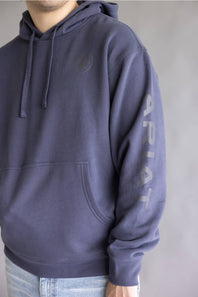 A person wearing a dark slate blue hoodie with a tonal chest logo and vertical 'ARIAT' text printed on the left sleeve, paired with light blue jeans against a neutral gray background.