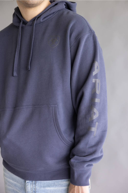 A person wearing a dark slate blue hoodie with a tonal chest logo and vertical 'ARIAT' text printed on the left sleeve, paired with light blue jeans against a neutral gray background.