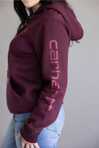 A person wearing a maroon Carhartt hoodie with a large tonal pink Carhartt logo printed vertically down the right sleeve, paired with light blue jeans against a plain gray background.