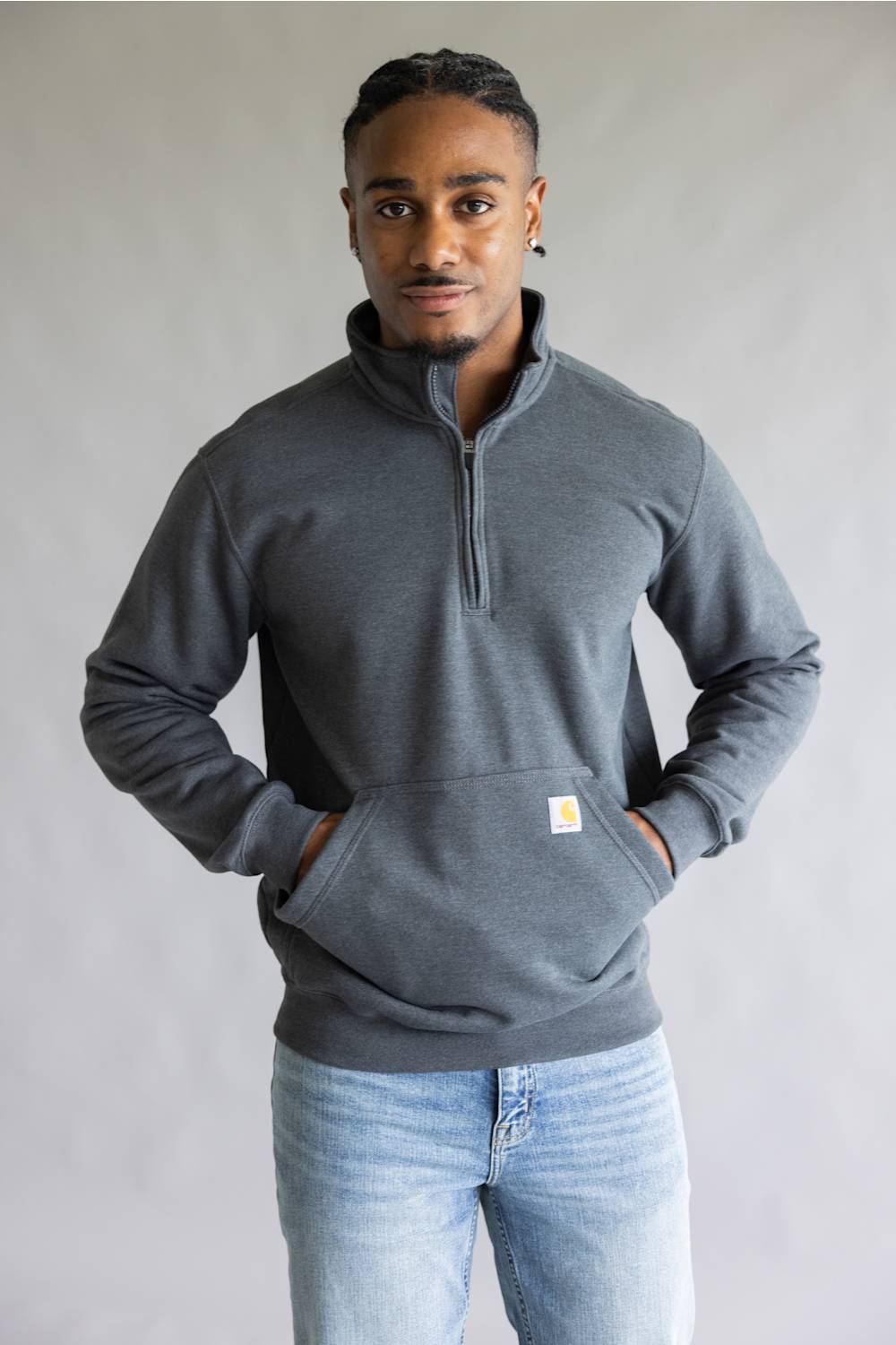 A man wearing a dark gray quarter-zip pullover sweatshirt with a small Carhartt logo patch on the front pocket, paired with light blue jeans, standing against a neutral gray background.