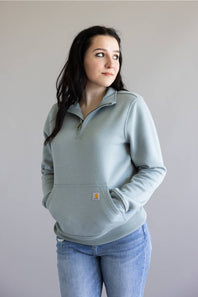 A woman wearing a light sage green quarter-zip sweatshirt with a kangaroo pocket and blue jeans, standing against a plain light gray background.
