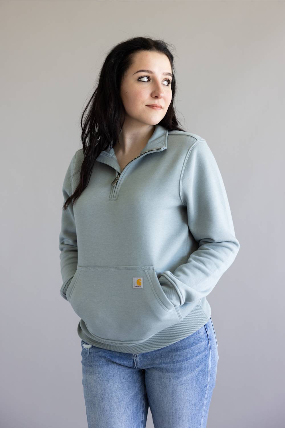 A woman wearing a light sage green quarter-zip sweatshirt with a kangaroo pocket and blue jeans, standing against a plain light gray background.
