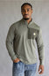 A man wearing a sage green ribbed quarter-zip pullover with a chest pocket and a small Carhartt logo, paired with blue jeans, standing against a neutral gray studio background.