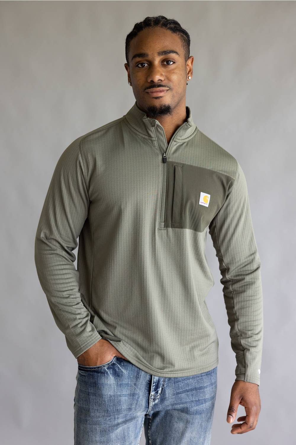 A man wearing a sage green ribbed quarter-zip pullover with a chest pocket and a small Carhartt logo, paired with blue jeans, standing against a neutral gray studio background.