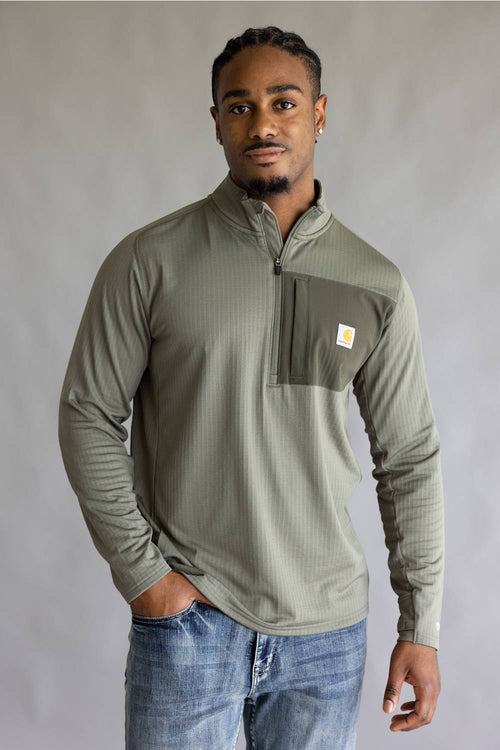 A man wearing a sage green ribbed quarter-zip pullover with a chest pocket and a small Carhartt logo, paired with blue jeans, standing against a neutral gray studio background.