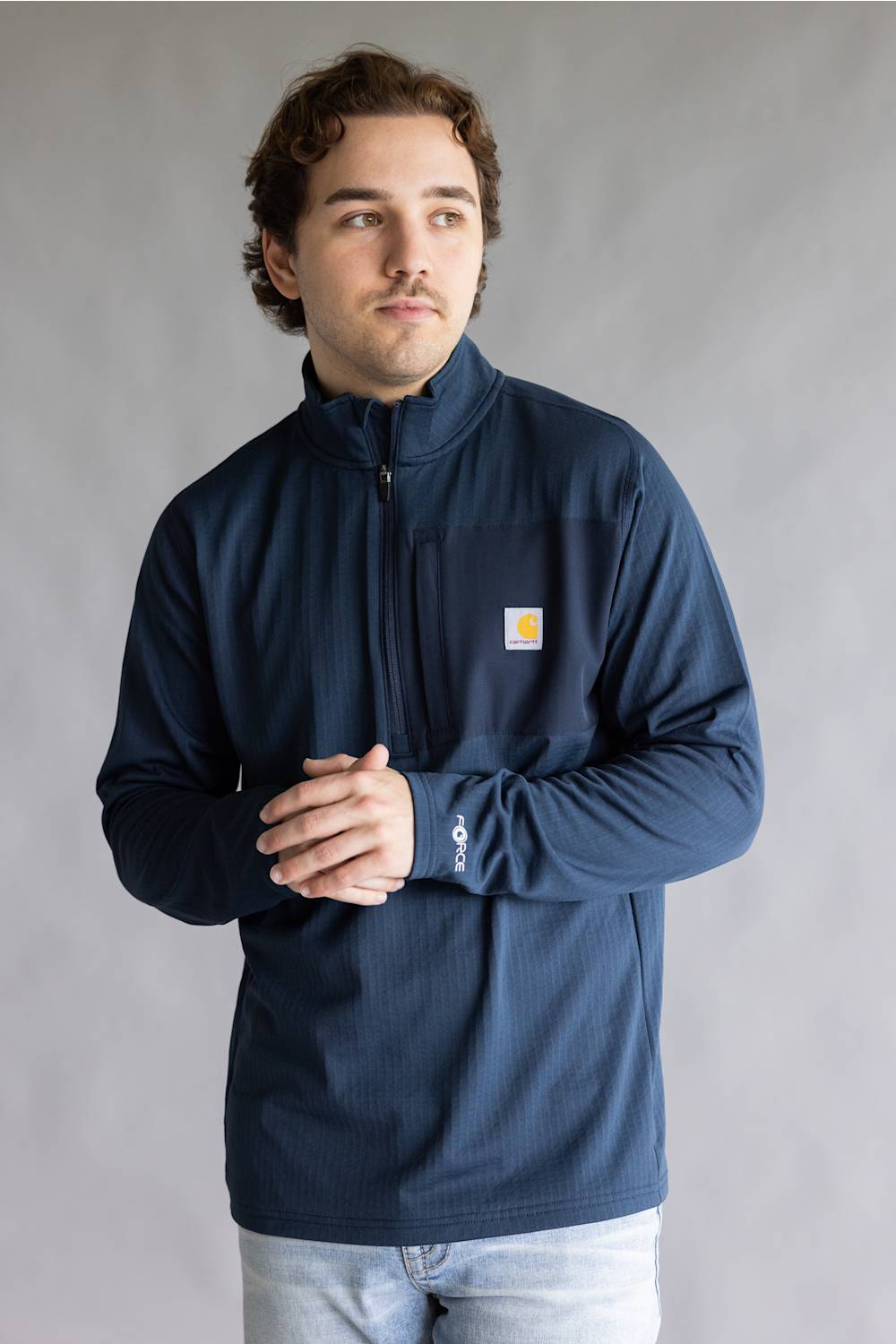 A man wearing a navy blue quarter-zip fleece jacket with a small yellow Carhartt logo on the left chest, standing against a neutral gray background.