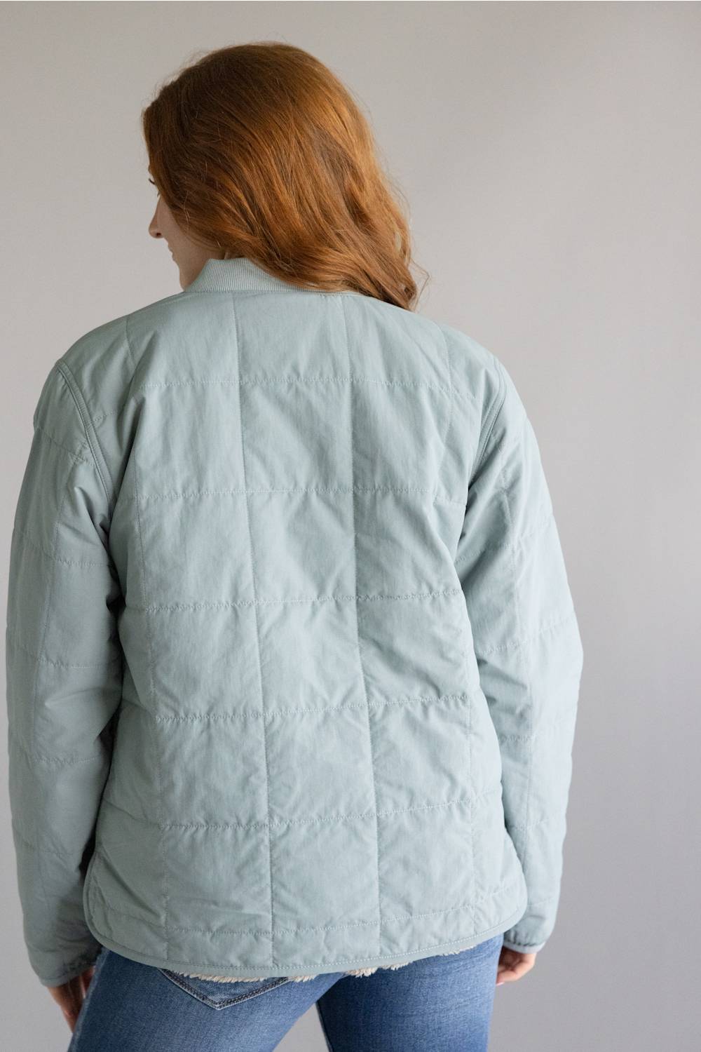 A person shown from the back wearing a pale sage green quilted jacket and blue jeans against a plain light gray background.