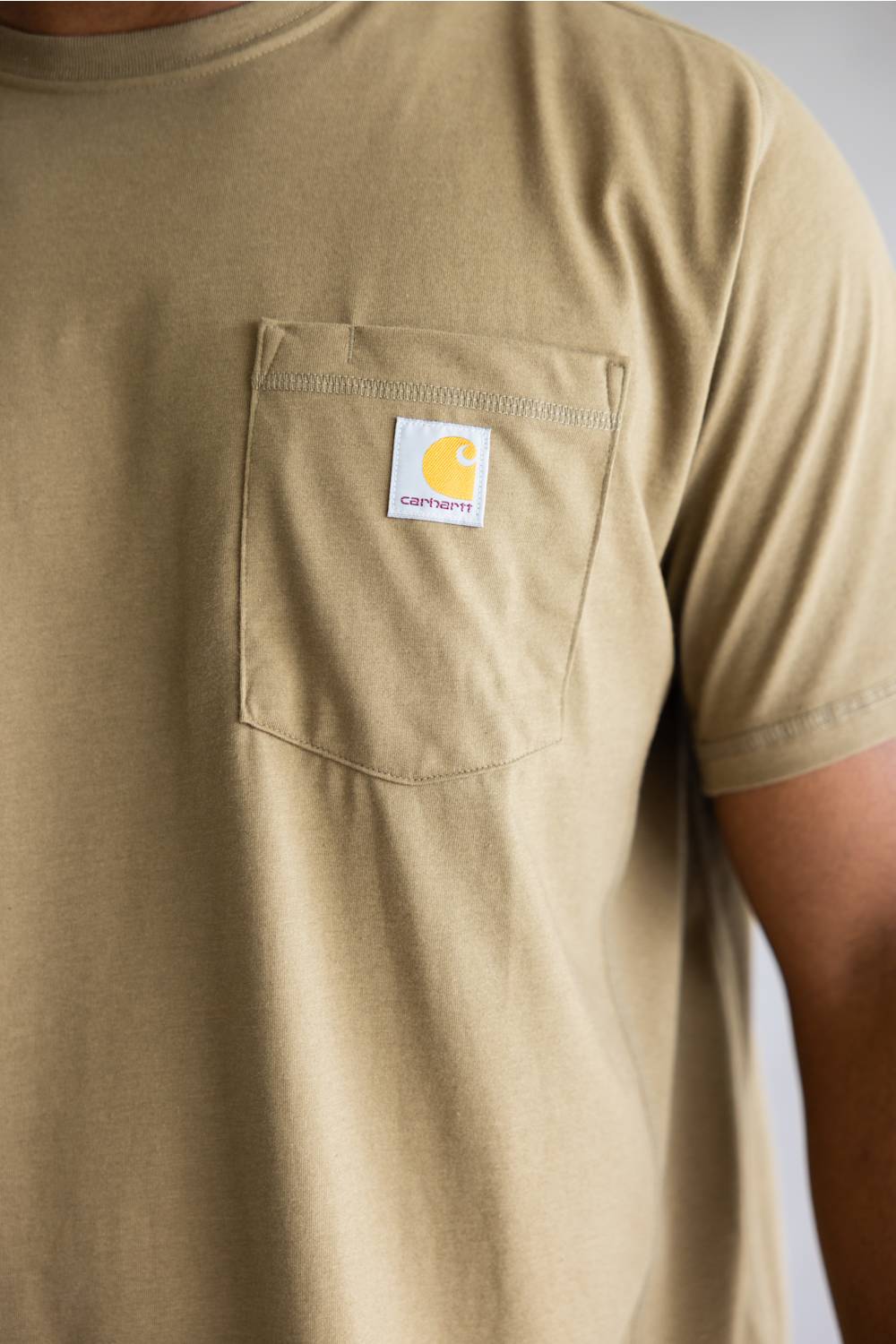 Close-up of a person wearing a tan short-sleeve pocket t-shirt with a white Carhartt logo patch on the chest pocket.