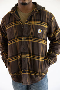 A man wearing a brown and yellow plaid hooded button-up overshirt by Carhartt over a green t-shirt and blue jeans, photographed against a plain white background.