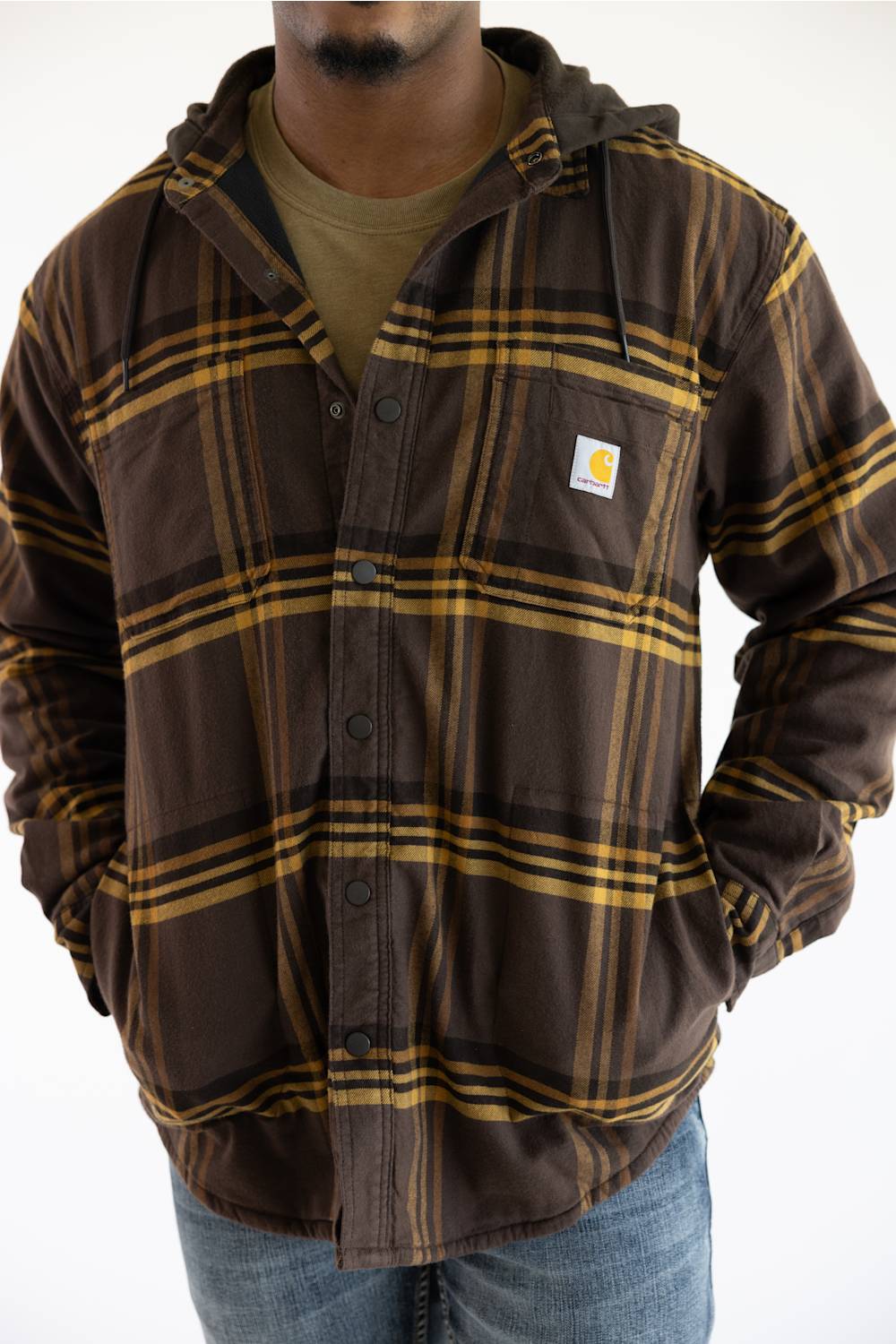 A man wearing a brown and yellow plaid hooded button-up overshirt by Carhartt over a green t-shirt and blue jeans, photographed against a plain white background.