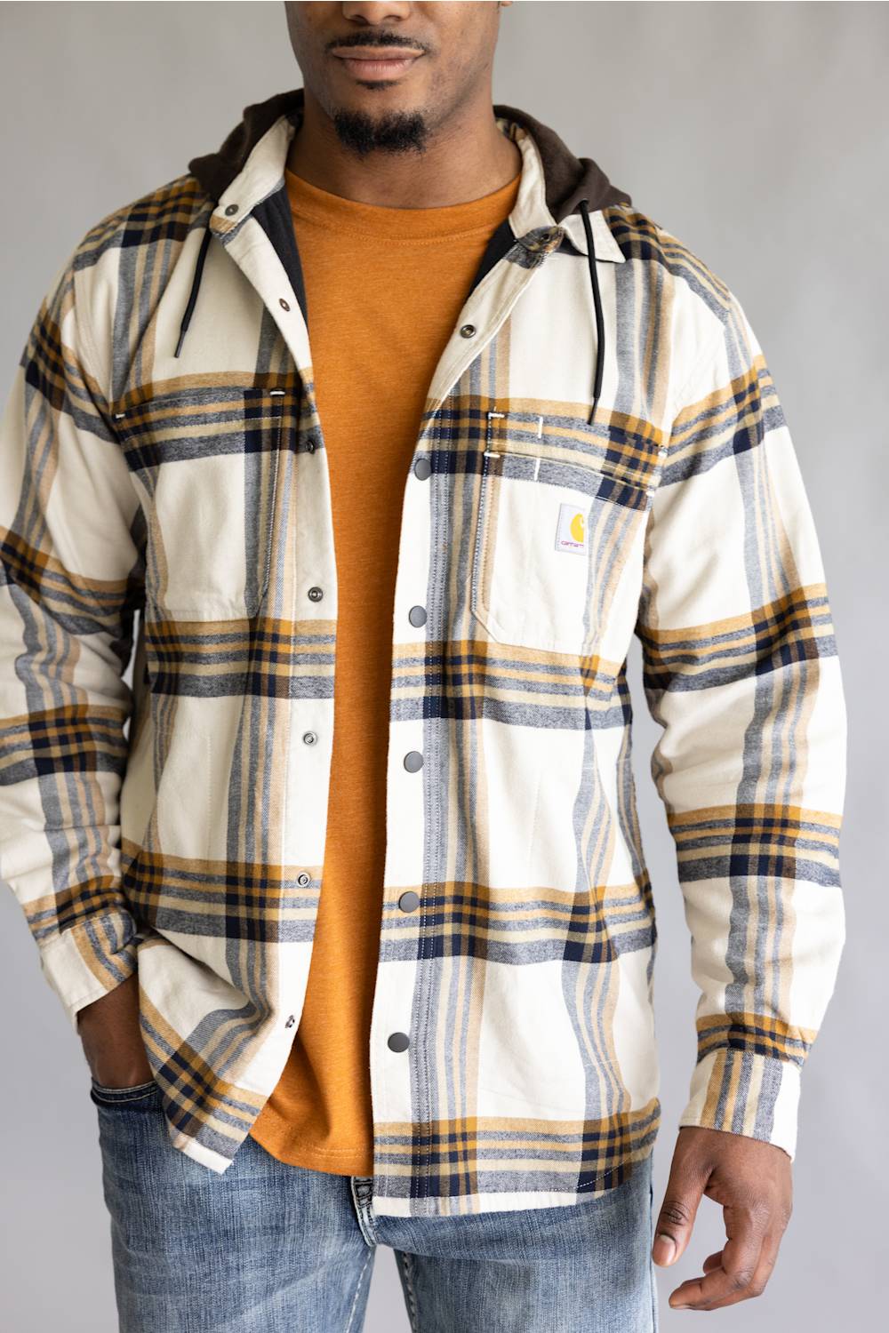 A man wearing a hooded plaid flannel shirt by Carhartt over an orange t-shirt and blue jeans, shown from the chin down against a neutral gray background.