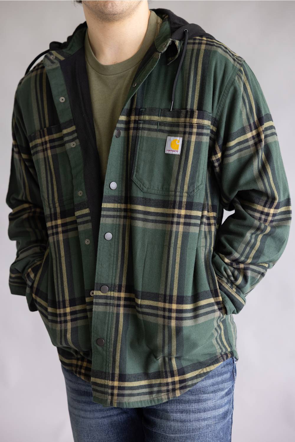 A man wearing a green Carhartt hooded flannel shirt jacket with a tan and black plaid pattern, layered over an olive green t-shirt and blue jeans.