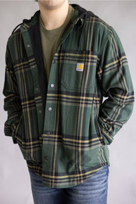 A man wearing a green Carhartt hooded flannel shirt jacket with a tan and black plaid pattern, layered over an olive green t-shirt and blue jeans.