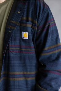 Close-up of a Carhartt striped overshirt in navy blue with red and yellow stripes, worn over a green shirt.