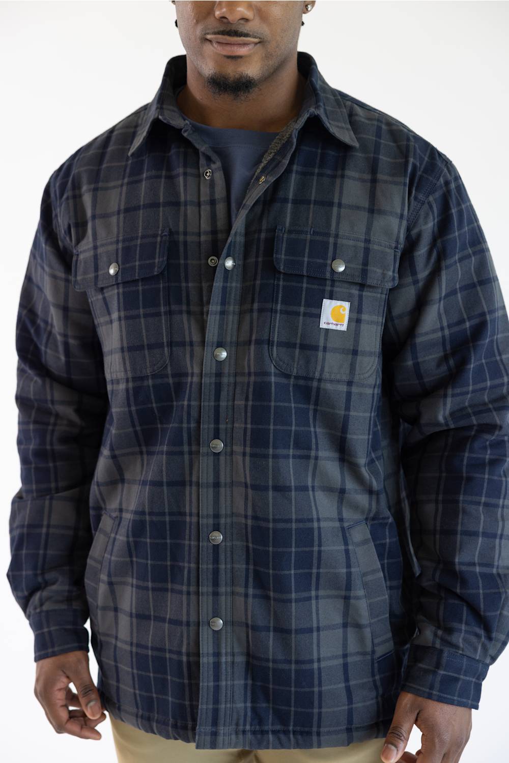 A man wearing a navy blue and gray plaid button-up shirt jacket with two chest pockets and a visible Carhartt logo patch on the left chest, photographed against a plain white background.