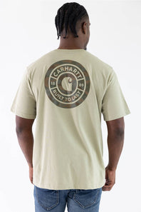 A man viewed from behind wearing a light beige Carhartt t-shirt with a large circular logo on the back, paired with blue jeans against a white studio background.